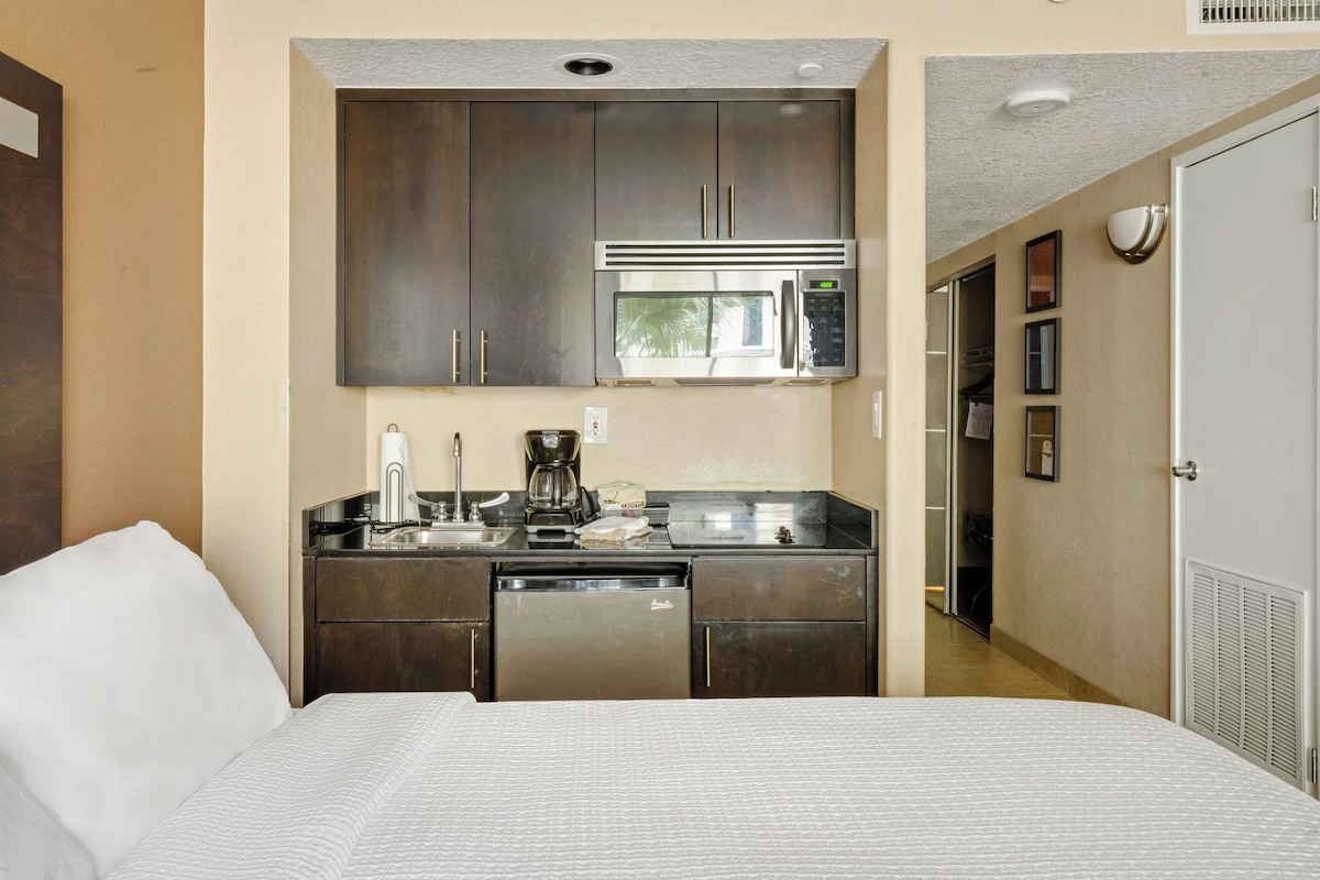 A compact hotel room with a kitchenette, a bed, and a door leading to a bathroom.