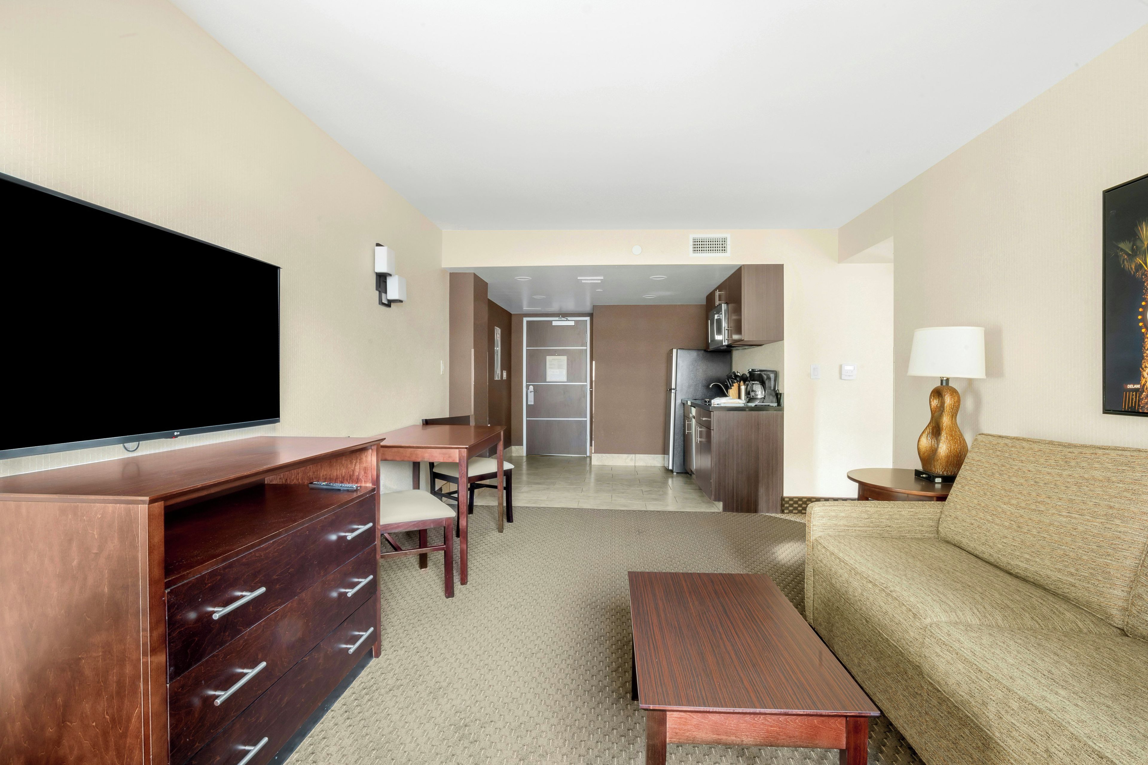 Habitación de hotel con televisor de pantalla plana, cómoda de madera, sofá, mesa de centro, mesa de comedor con sillas y kitchenette.
