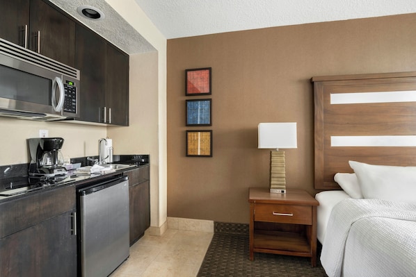 Una habitación de hotel con kitchenette, una cama, una mesita de noche con lámpara y una pared con tres cuadros enmarcados.