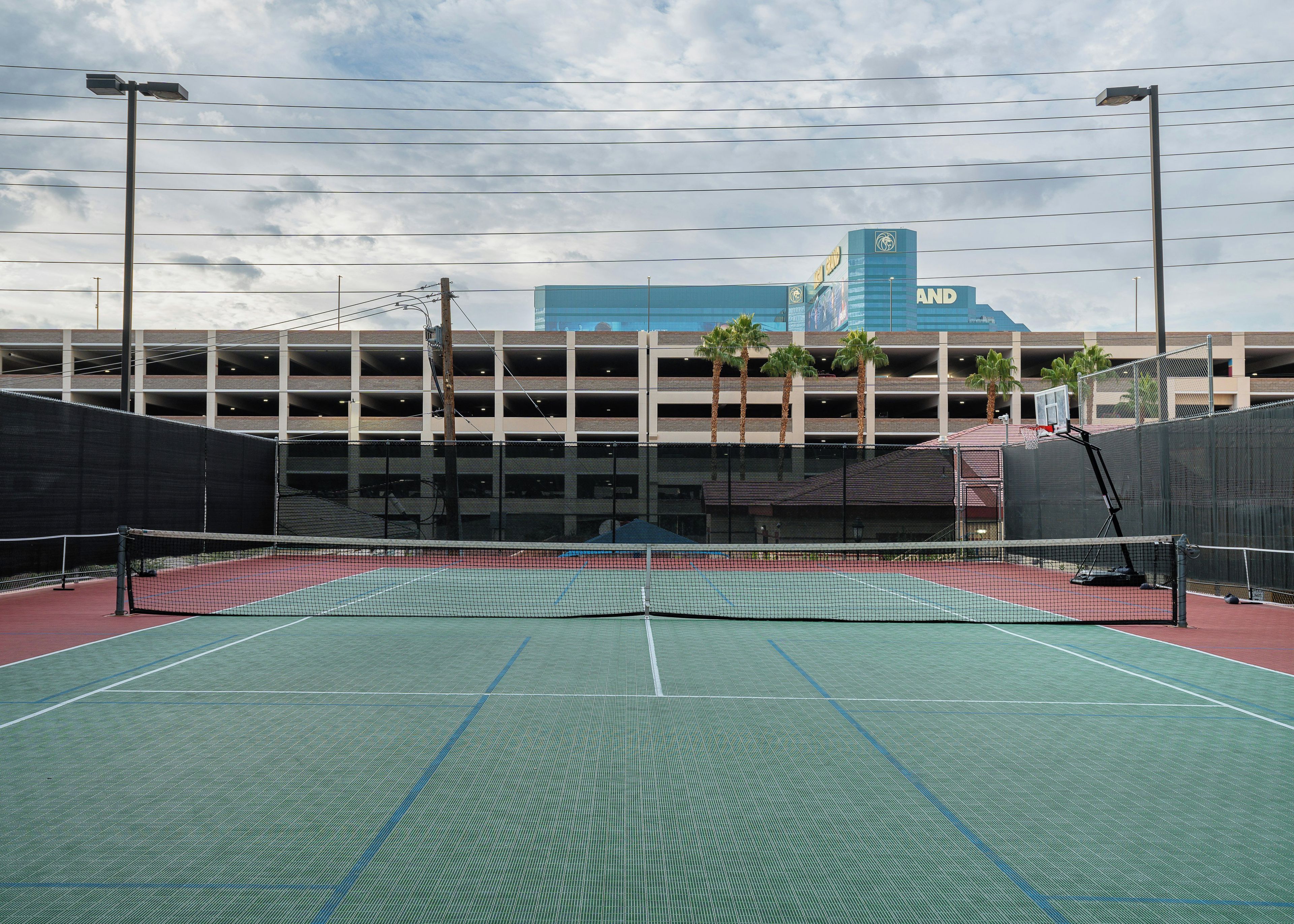 Una cancha de tenis exterior con red, rodeada por una valla, y un estacionamiento al fondo.