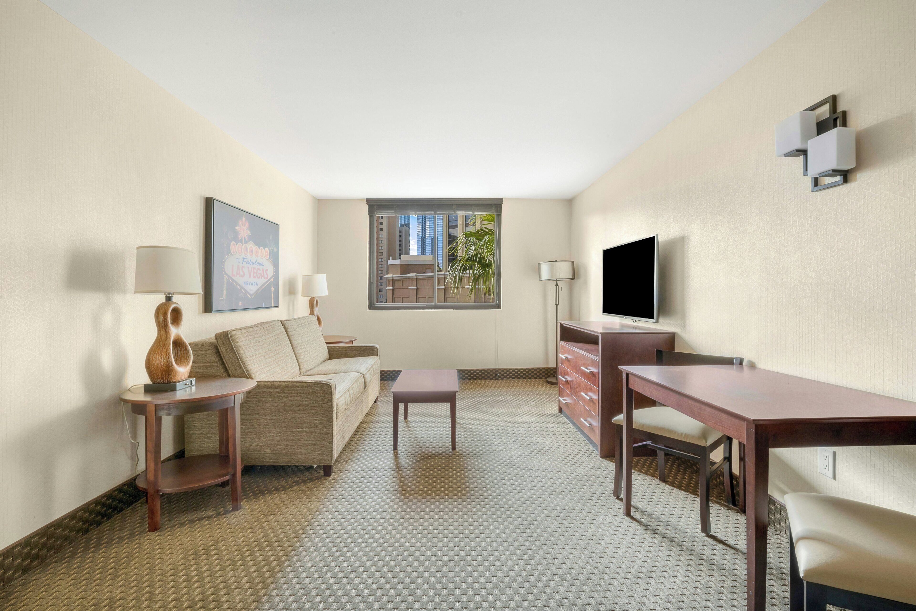 A hotel room with a sofa, a small table, a desk, a TV, and a window with a view of buildings.