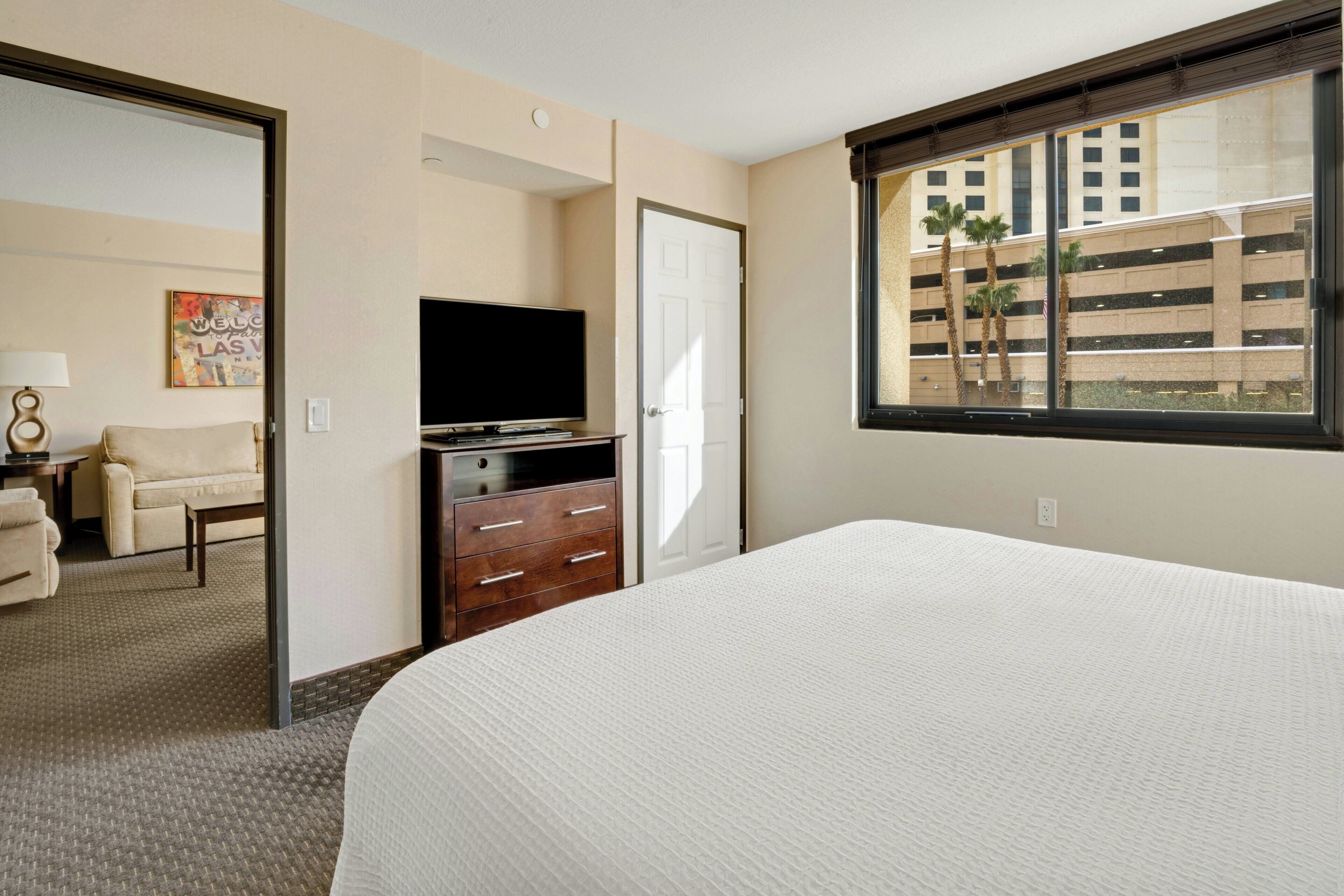 A hotel room with a bed, a dresser, a television, and a window with a view of a building and palm trees.
