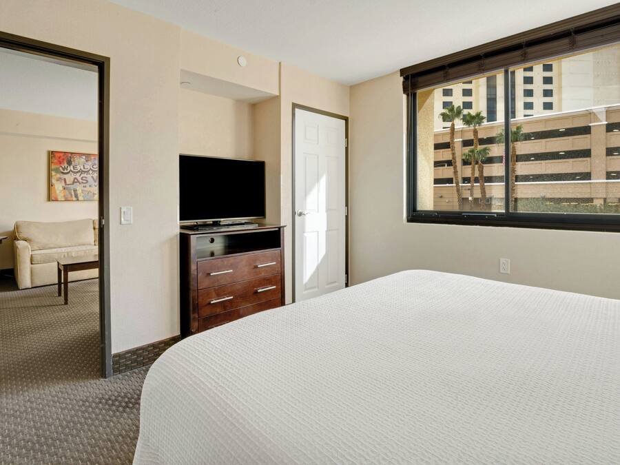 A hotel room with a bed, a dresser, a television, and a window with a view of a building and palm trees.