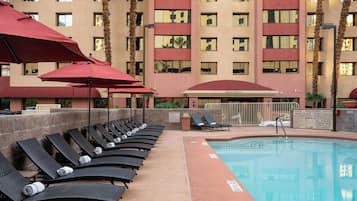 Uma área de piscina com espreguiçadeiras e guarda-sóis, um edifício de recepção e a fachada de um hotel com vários andares.