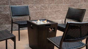A square outdoor seating area with a built-in fire feature, surrounded by chairs with woven backs.