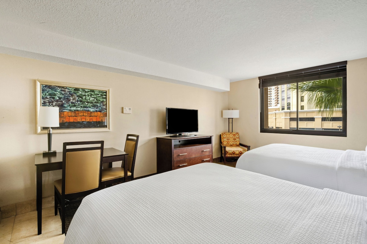 A hotel room with a bed, a desk with a chair, a television, a lamp, and a window with a view of buildings.