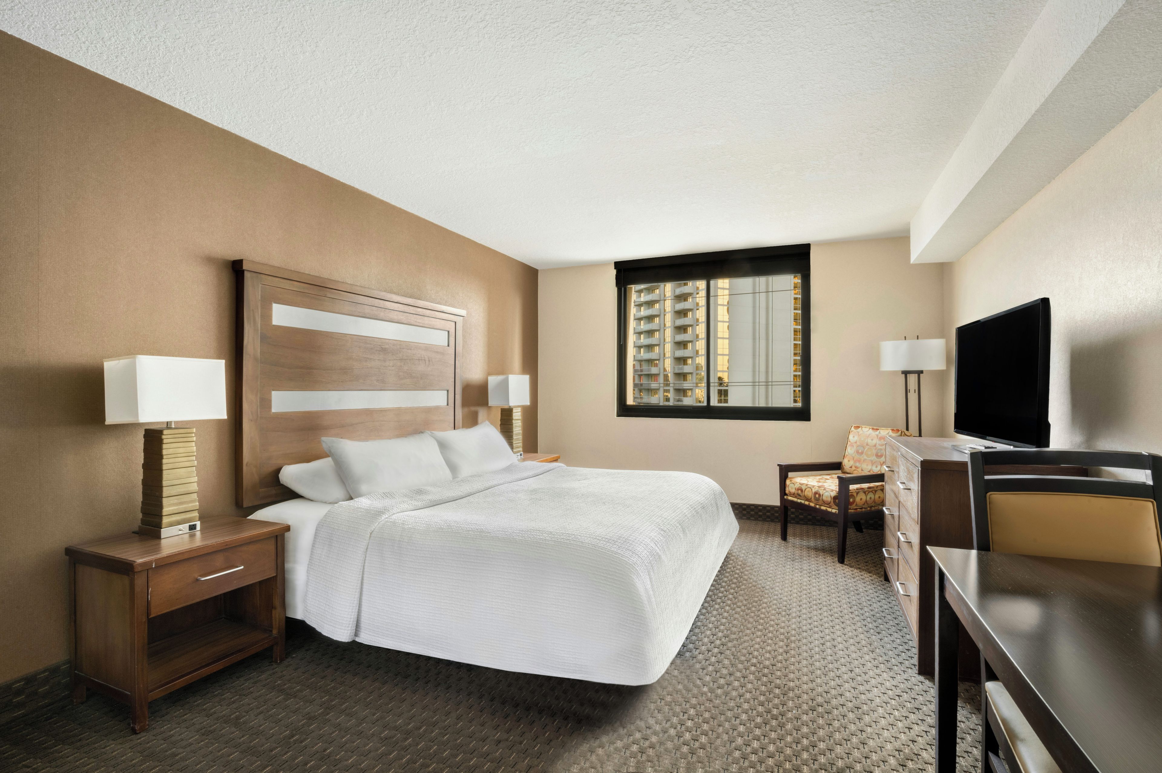 A hotel room with a bed, bedside tables, a desk, a chair, a television, and a window with a view of buildings.