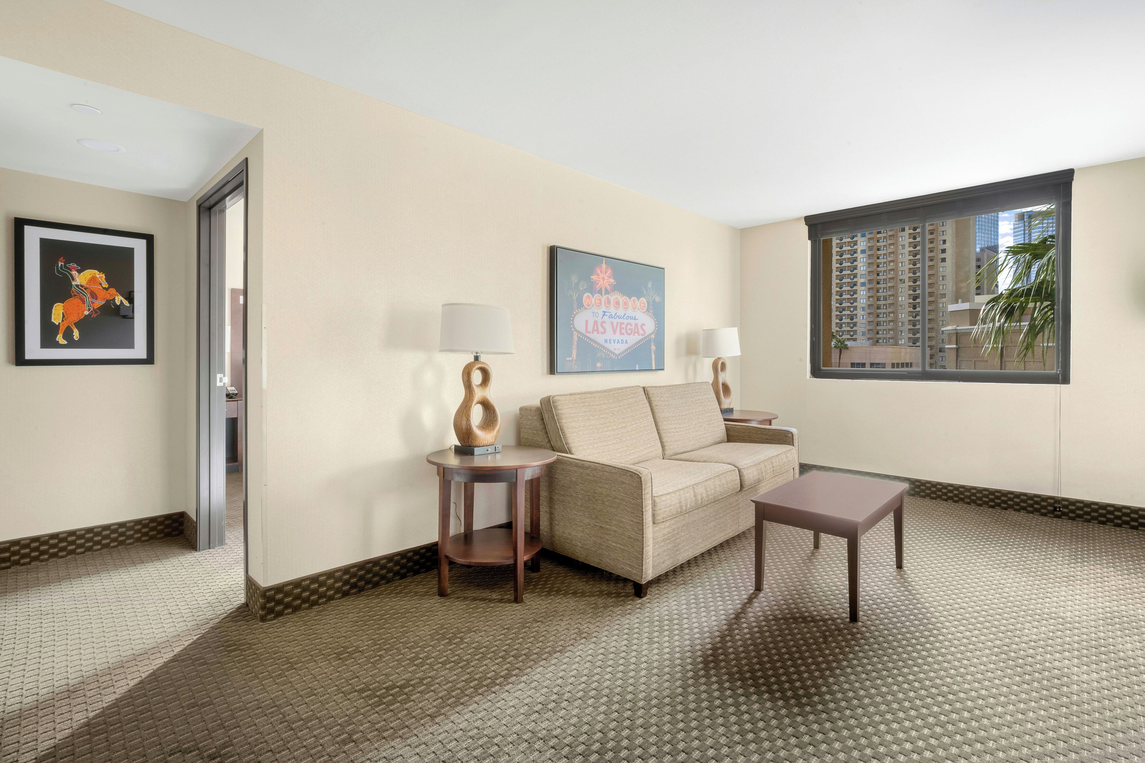 Habitación de hotel con sofá, una mesita, lámpara, un cuadro enmarco y ventana con vista a edificios.