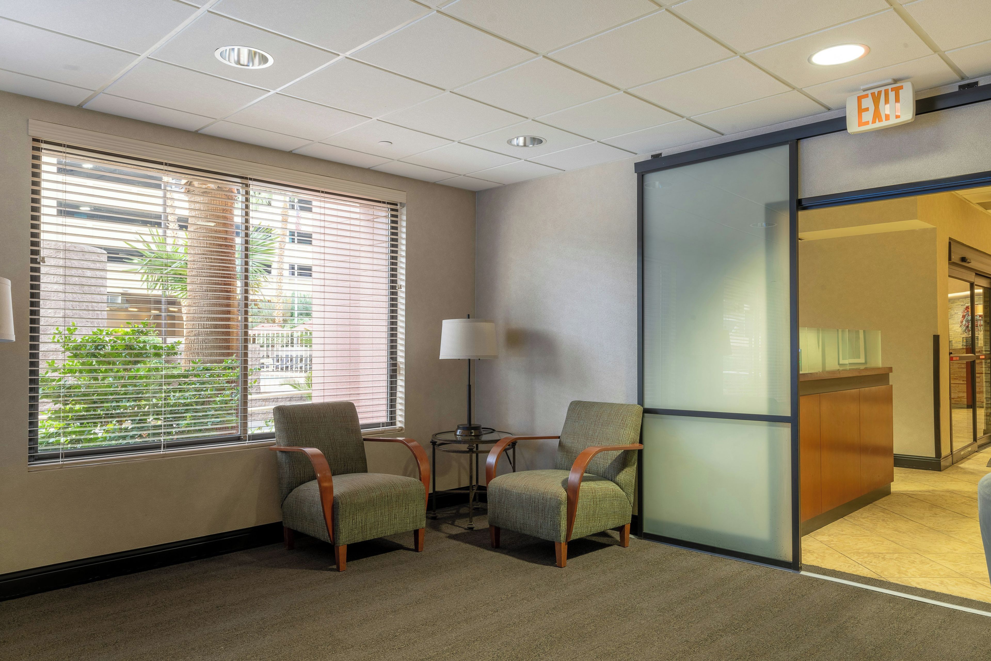 A hotel lobby with two armchairs, a lamp, and a glass partition with an exit sign.