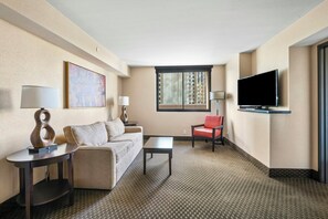 Habitación de hotel con sofá, una silla, una mesita de centro, televisor y ventana con persianas.