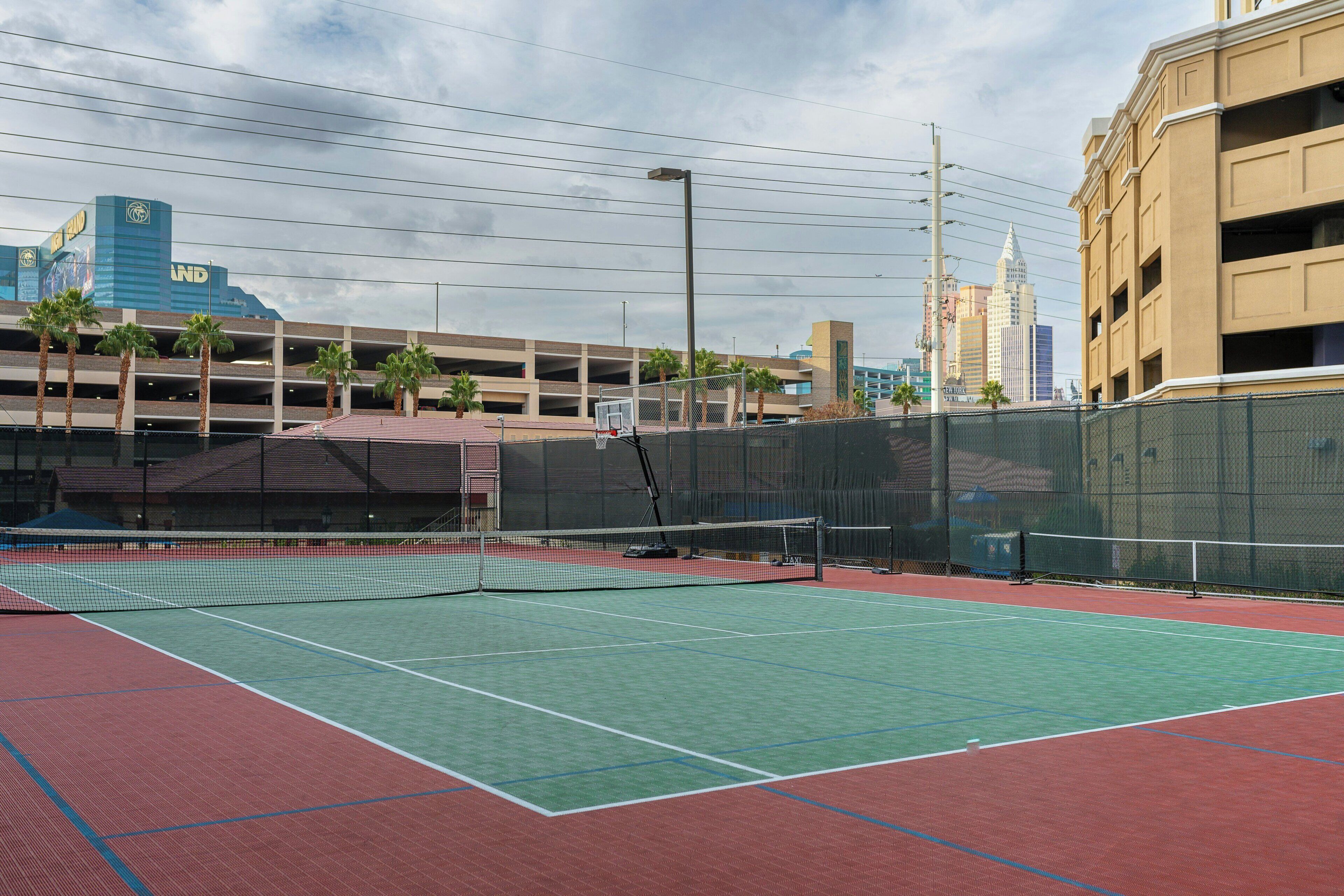 An outdoor tennis court with a net, surrounded by a fence, and a parking garage in the background.