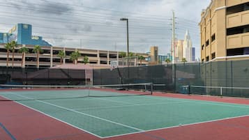 An outdoor tennis court with a net, surrounded by a fence, and a parking garage in the background.