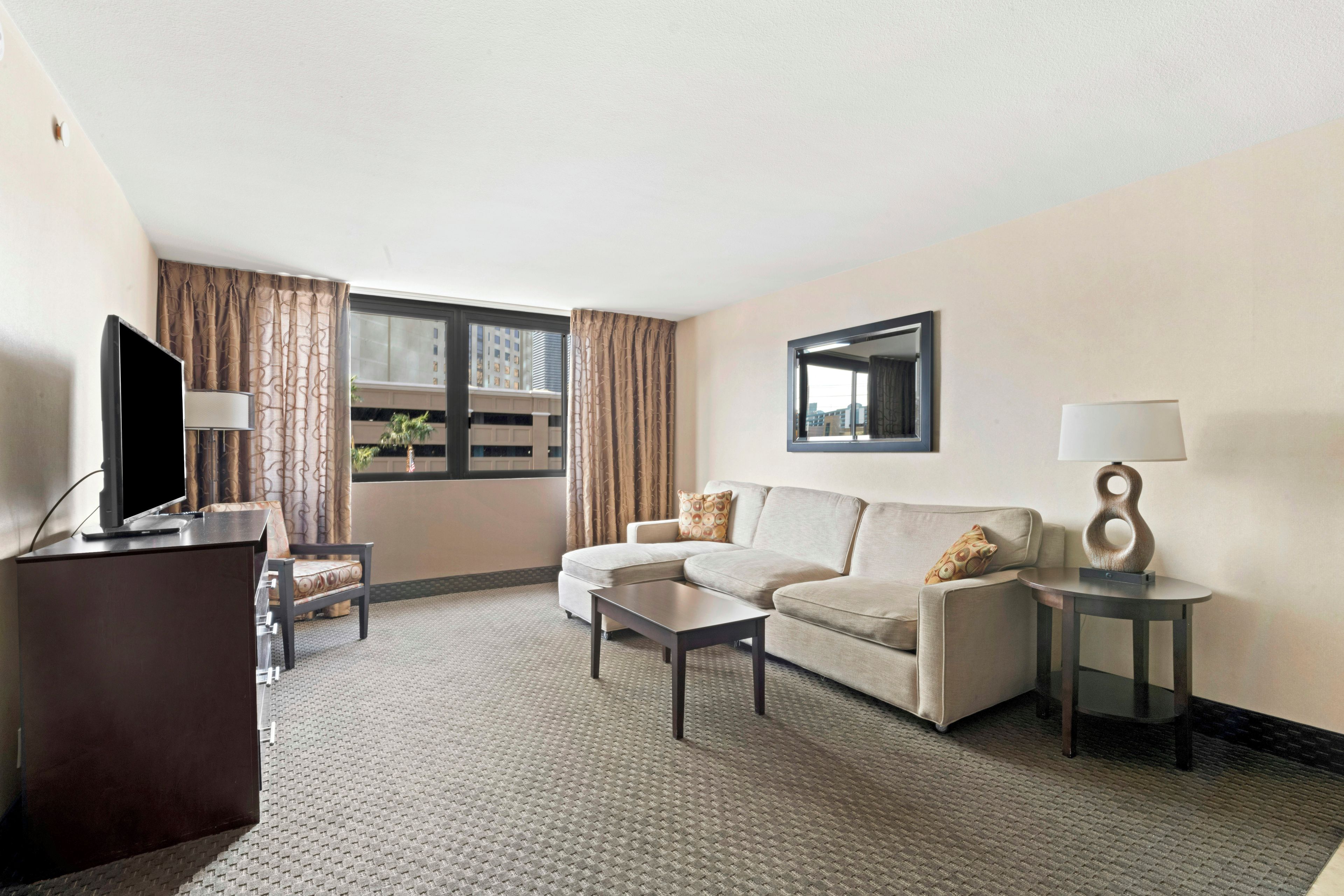 A hotel room with a sofa, a coffee table, a TV, a lamp, and a window with curtains.