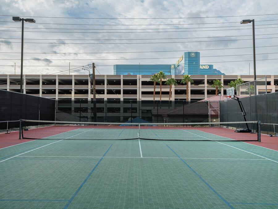 An outdoor tennis court with a net, surrounded by a fence, and a parking garage in the background.