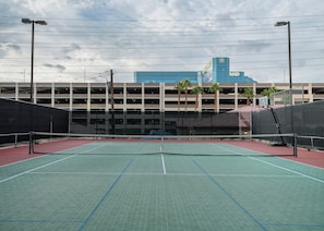 An outdoor tennis court with a net, surrounded by a fence, and a parking garage in the background. - The Carriage House (Las Vegas)