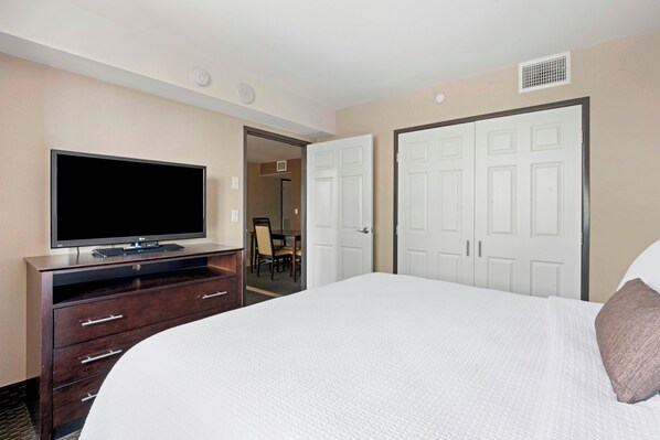 A hotel room with a bed, a TV on a wooden stand, a door, and a hallway leading to a dining area.