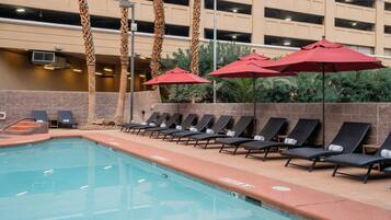 A pool area with lounge chairs and umbrellas, surrounded by palm trees and a building with multiple floors.