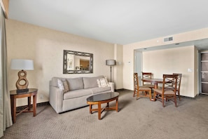 A hotel room with a sofa, a coffee table, a dining table with chairs, a lamp, and a mirror on the wall.