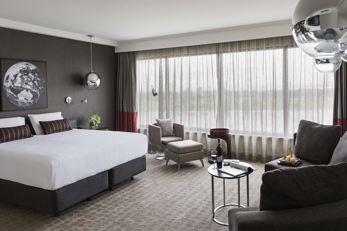 Una habitación de hotel moderna con una cama grande, una zona de estar con una silla y un sofá, una mesita con una copa de vino y una botella, y un ventanal con cortinas.
