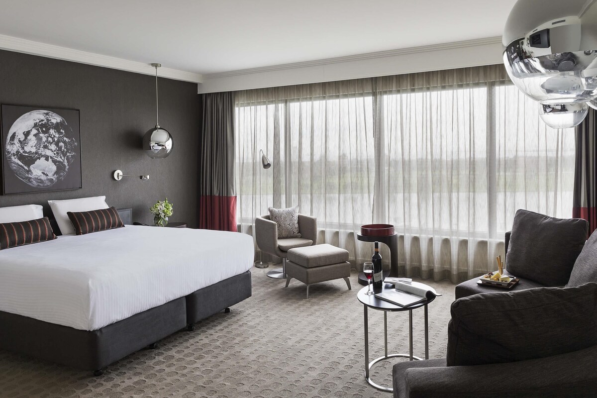 Una habitación de hotel moderna con una cama grande, una zona de estar con una silla y un sofá, una mesita con una copa de vino y una botella, y un ventanal con cortinas.