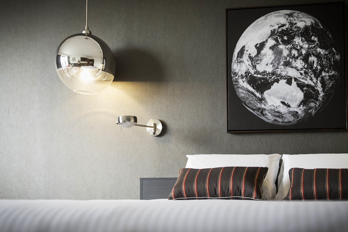 A bedroom with a bed, pillows, a wall-mounted light fixture, and a framed picture of the Earth.