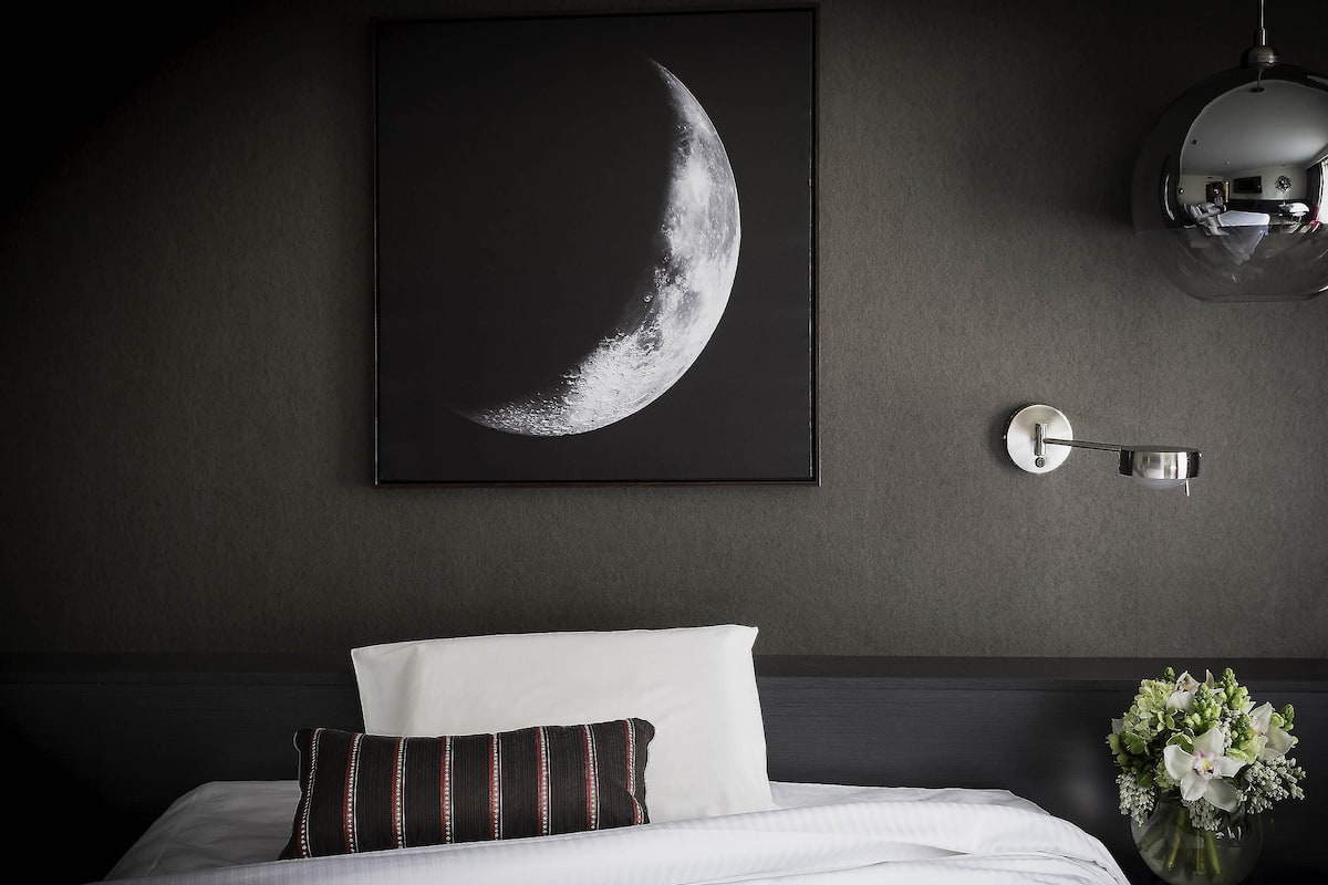 A bedroom with a bed, a wall-mounted picture of a crescent moon, a wall sconce, and a vase with flowers.