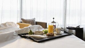 Un plateau de petit-déjeuner bien présenté, avec une cafetière, un verre de jus d’orange, un bol de céréales, une poire et un croissant, posé sur une table de chevet dans une chambre d’hôtel.