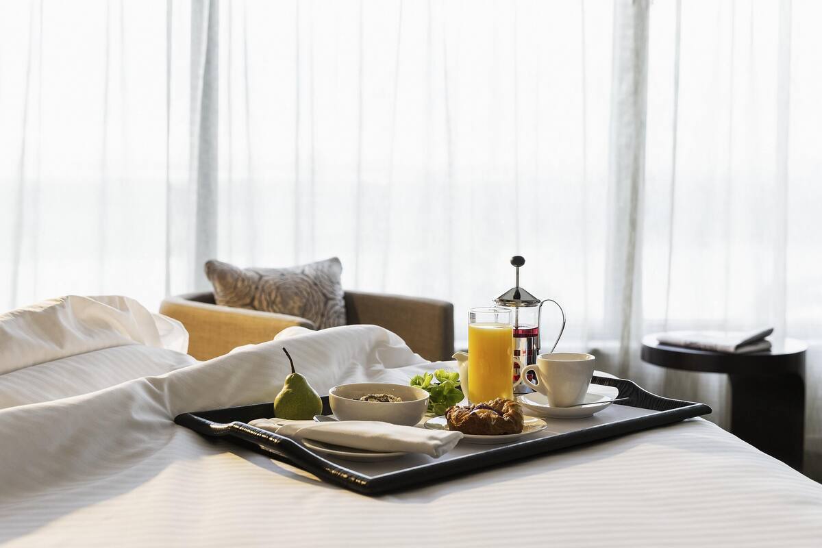 A neatly made breakfast tray with a coffee pot, a glass of orange juice, a bowl of cereal, a pear, and a croissant on a bed in a hotel room.