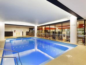 An indoor swimming pool with exercise equipment, a reception area, and a view of the outdoors through large windows.