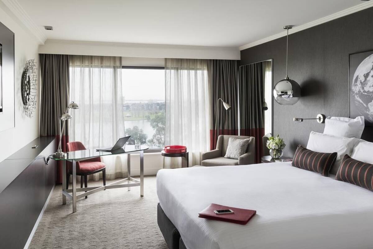 A hotel room with a large bed, a desk with a laptop, a chair, a small table with a red bowl, and a view of the city through the window.