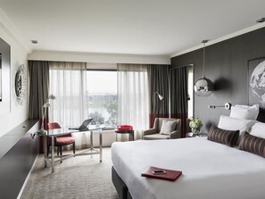 A hotel room with a large bed, a desk with a laptop, a chair, a small table with a red bowl, and a view of the city through the window.