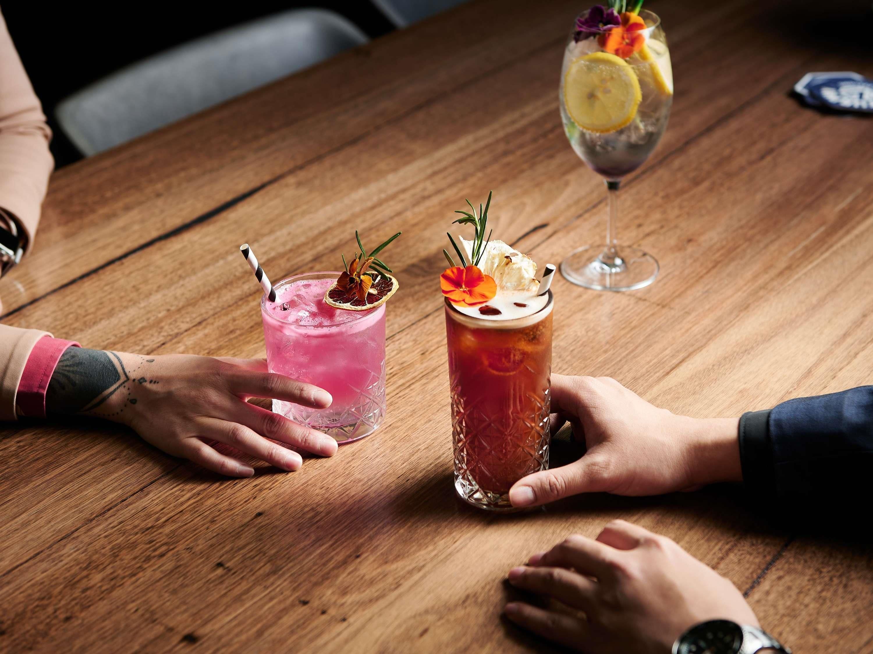 A wooden table with two drinks, one pink and one red, garnished with flowers and herbs.