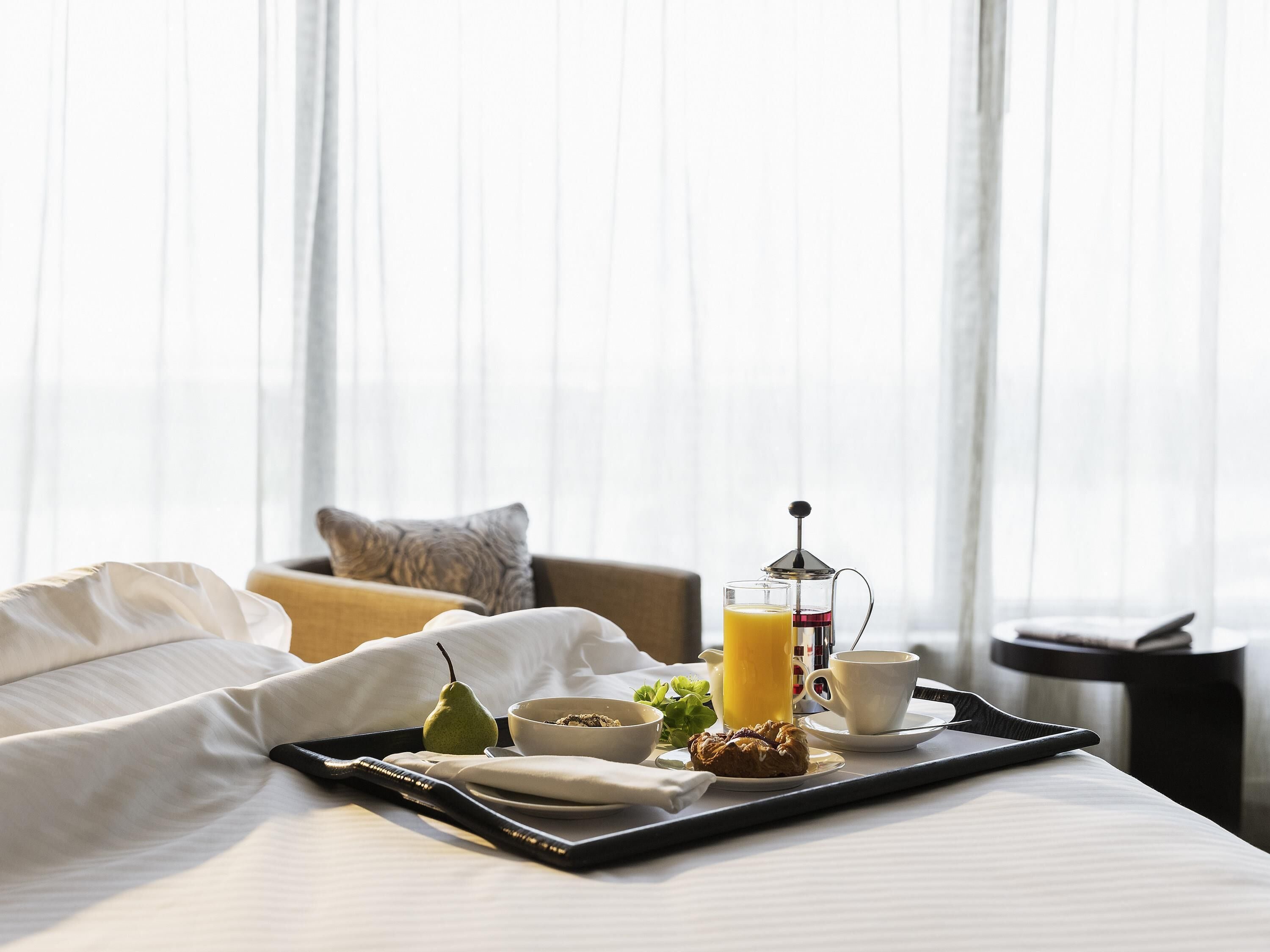 A neatly made bed with a tray of breakfast items including a pear, a croissant, a glass of orange juice, a cup of coffee, and a French press.