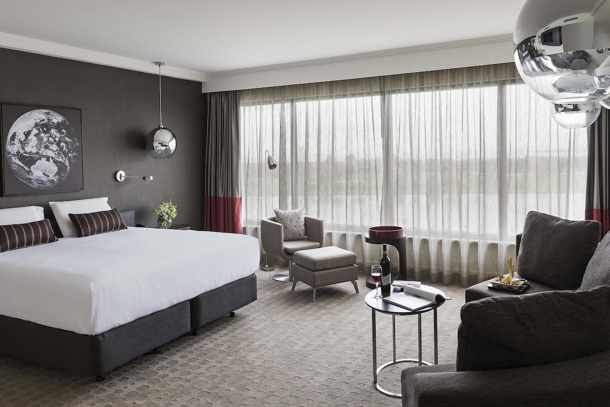 A modern hotel room with a large bed, a seating area with a chair and sofa, a small table with a wine glass and bottle, and a large window with curtains.
