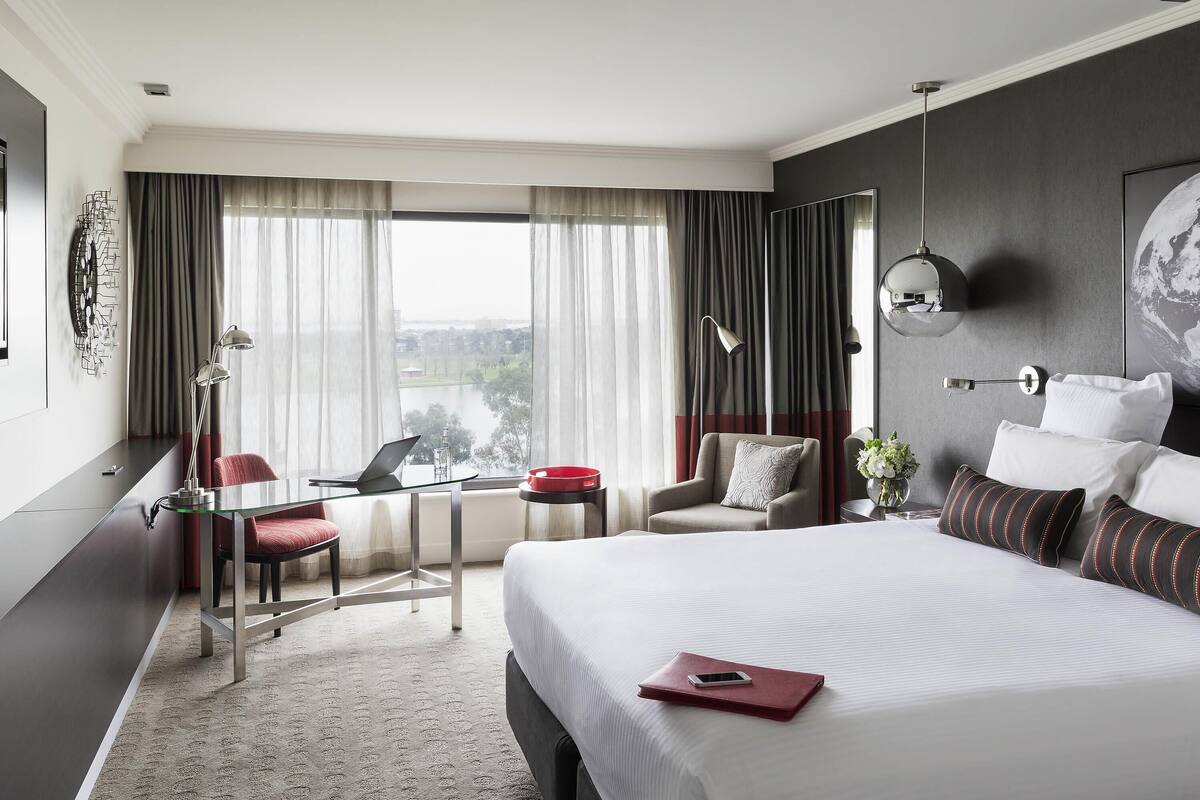 A modern hotel room with a large bed, a desk with a laptop, a chair, and a view of a cityscape through the window.