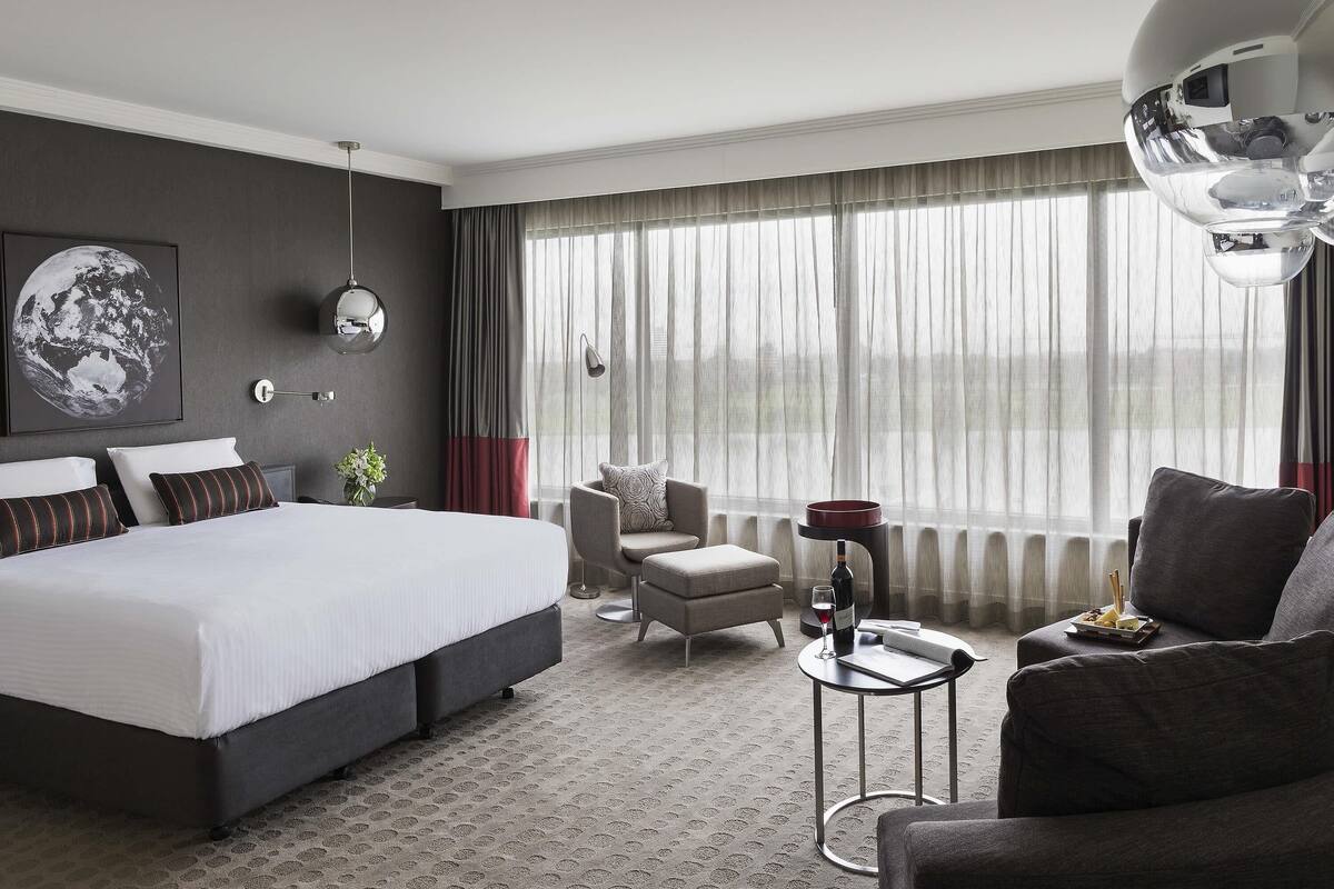 A modern hotel room with a large bed, a seating area with a chair and sofa, a small table with a wine glass and bottle, and a large window with curtains.