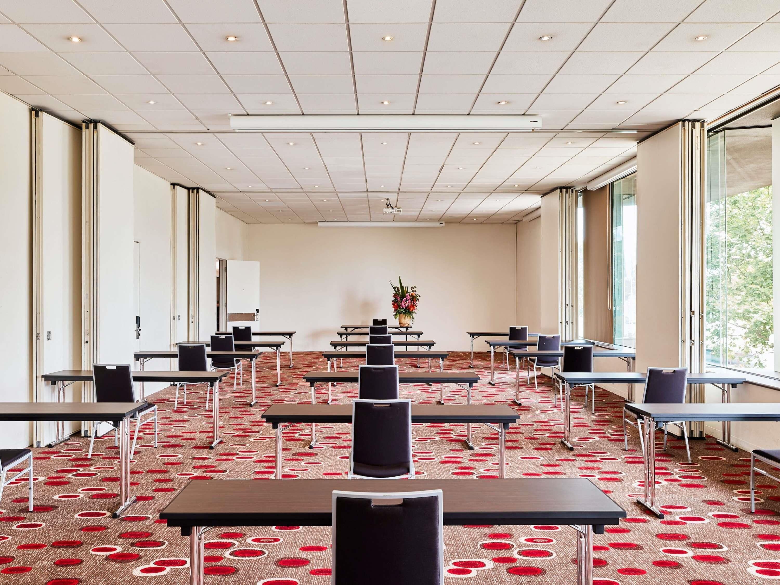 A conference room with tables and chairs, a projector screen, and a large window with a view of trees.