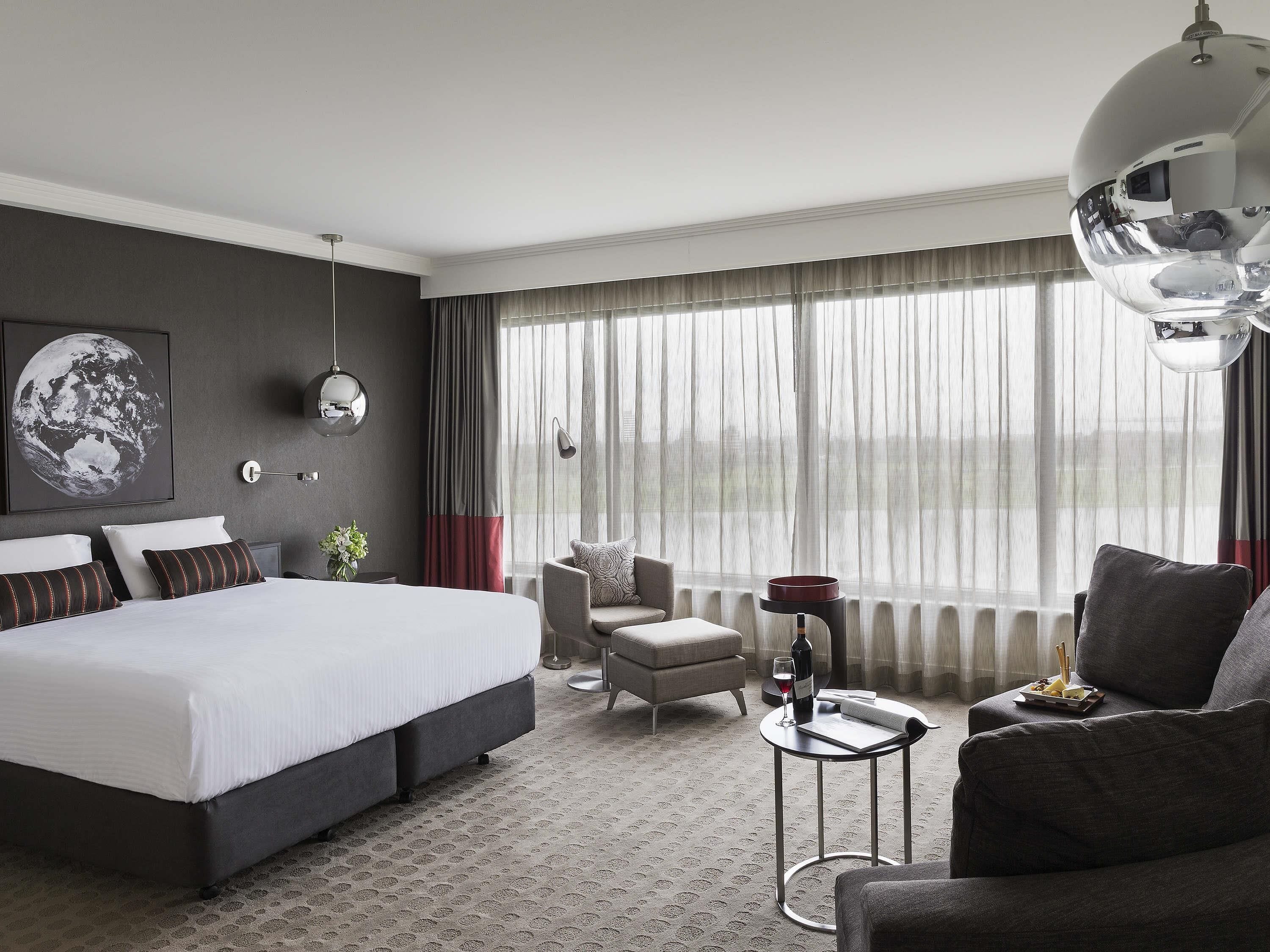 A modern hotel room with a large bed, a seating area with a chair and sofa, a small table with a wine glass and bottle, and a large window with curtains.