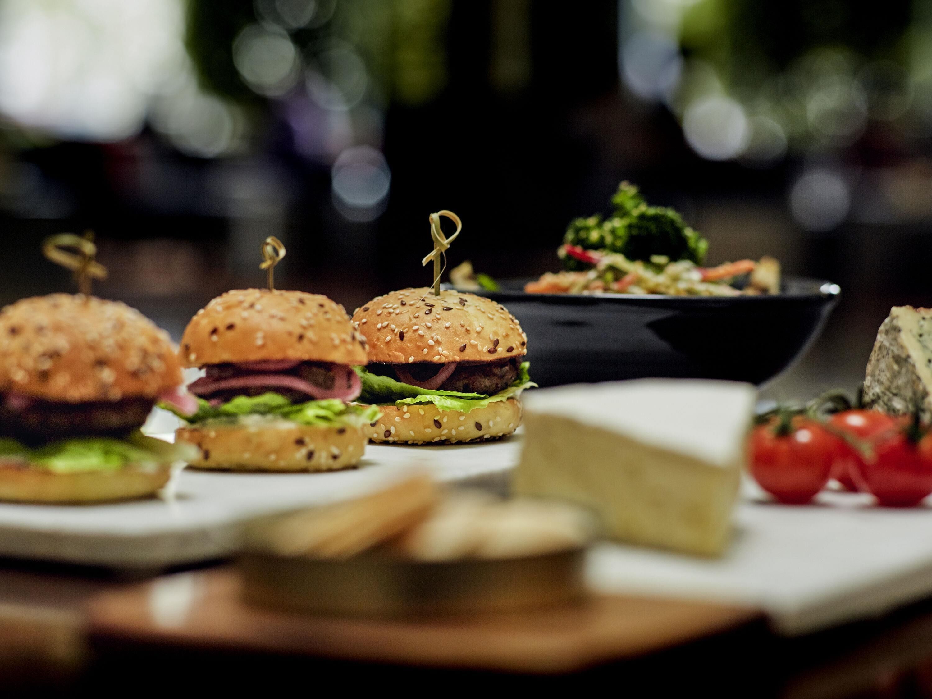Ein Tisch mit drei Burgern, einer Salat-Schüssel, einem Käse-Stück und einigen Tomaten.