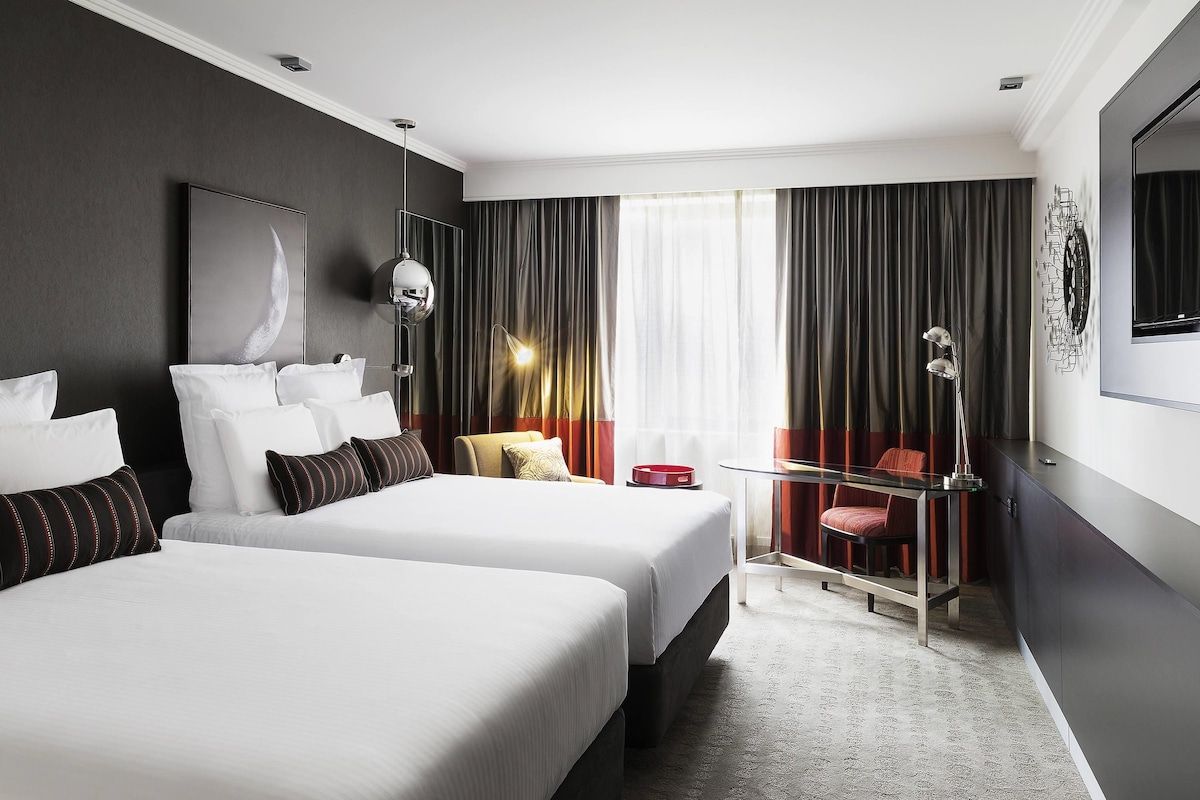 A hotel room with two beds, a desk, a chair, a TV, and a large window with curtains.
