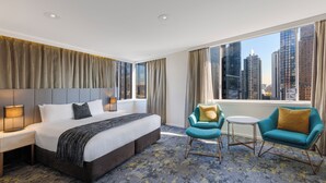Habitación de hotel con una cama grande, una mesita de noche, una silla, una pequeña mesa y vistas a edificios altos.