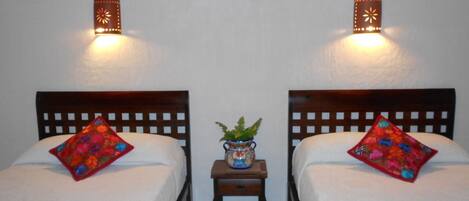 A room with two beds, each with a colorful striped bedspread and a floral pillow. There are wall-mounted lamps on either side of the beds, and a small table with a plant in the center.
