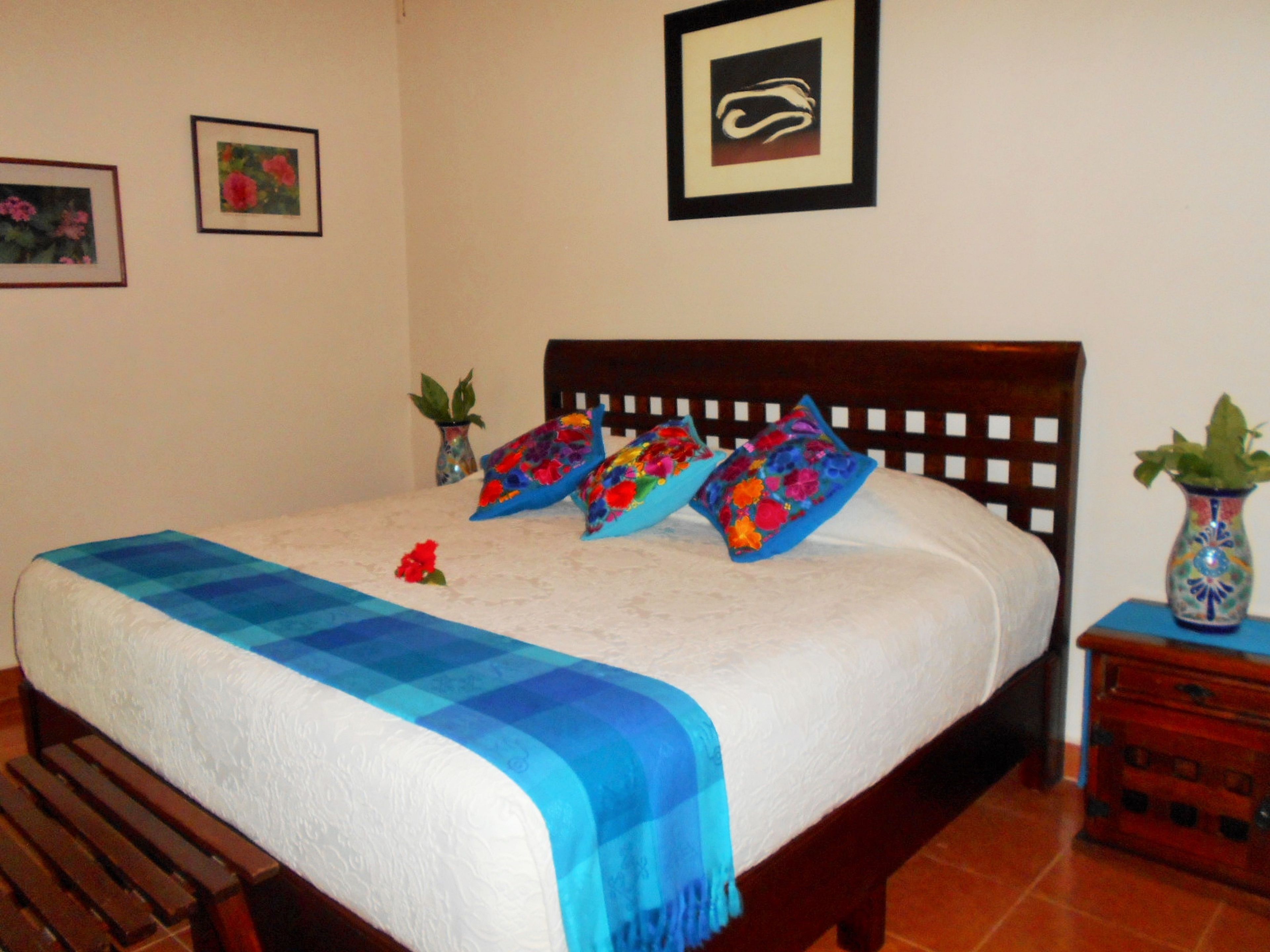 Una cama bien hecha con un edredón azul y blanco a cuadros, dos almohadas coloridas y una sola flor roja sobre la cama.