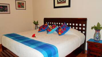 Una cama bien hecha con un edredón azul y blanco a cuadros, dos almohadas coloridas y una sola flor roja sobre la cama.
