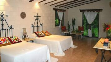 Una habitación con dos camas, una mesita, una silla y un ventanal con cortinas.