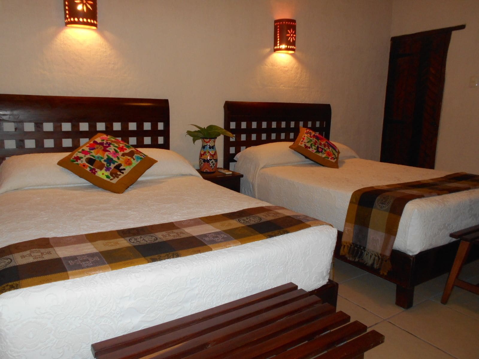 Habitación de hotel con dos camas, cada una con cabecero de madera, sábanas blancas y almohadas decorativas. Hay apliques de pared que proporcionan iluminación ambiental.