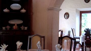 A dining room with a table set for four, featuring white plates with a monogram, clear glassware, and white napkin arrangements. A wooden china cabinet with various dishes is visible in the background.