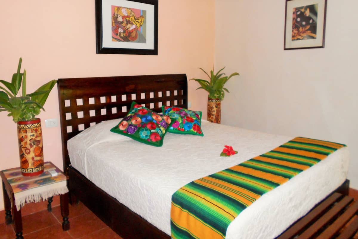 A neatly made bed with colorful pillows, a wooden headboard, and a striped blanket. There are two potted plants on either side of the bed.