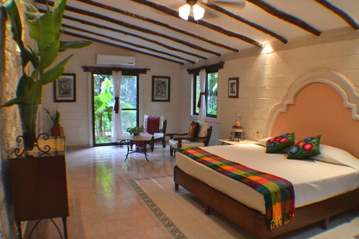 A bedroom with a large bed, a ceiling fan, a window with curtains, and a plant.