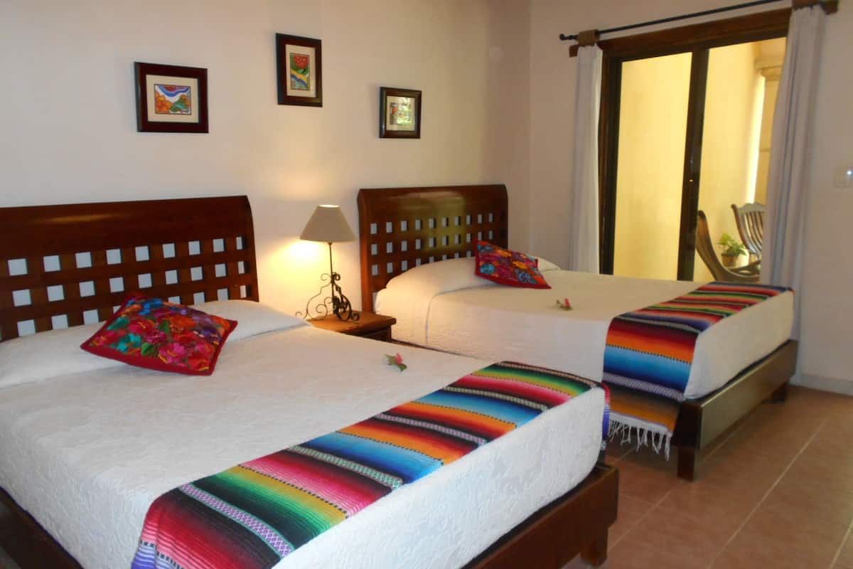 A hotel room with two beds, each with a colorful blanket and a pillow. There is a nightstand with a lamp between the beds. The room has a wooden headboard and a chair near the window.